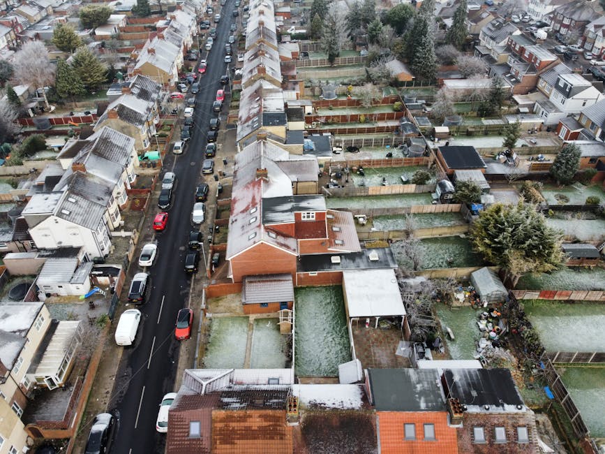 An aerial view of a residential neighborhood in Harlesden, showing closely packed terraced houses with varied roof styles, some covered in light snow or frost, alongside narrow streets lined with park