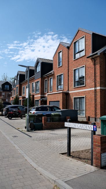 A modern residential street scene showing a multi-story brick building with large windows and pitched roofs, under a bright blue sky with some scattered clouds. In front of the building, there is a pa